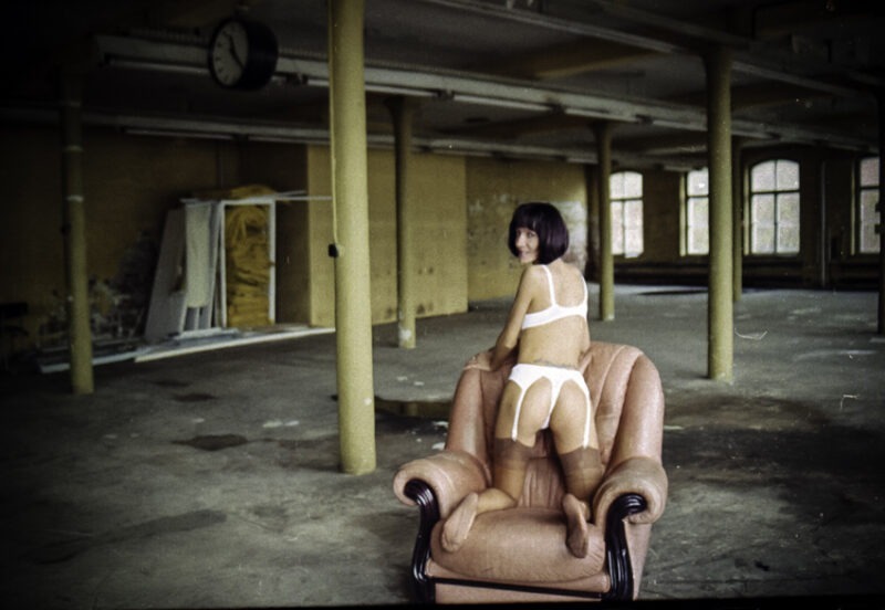 Sessel in der alten Fabrik. Beat kniet auf dem Sessel. Man sieht ihre Strumpf bestrumpften Füße. Sie trägt helle Nahtstrümpfe.