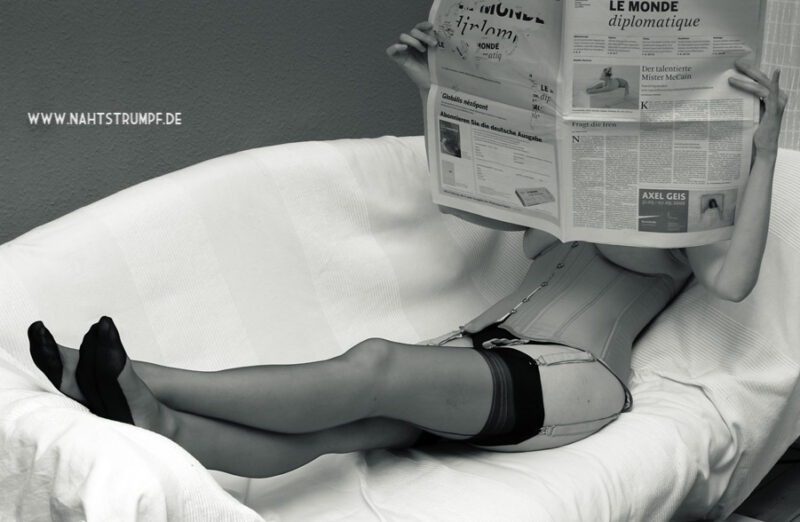 Eine Frau liest in Damenstrümpfen und Korsett gekleidet eine Tageszeitung. Dabei liegt sie auf einem Sofa.