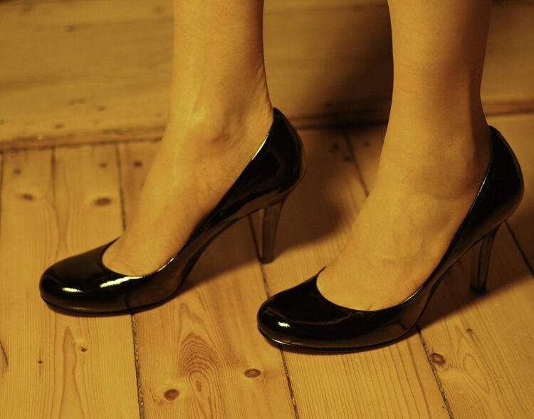 Nahaufnahme von Füßen in Schwarzen High Heels und hellen, feinen nahtlosen Strümpfen. Die Pumps sind aus Patentleder - also glänzend. Das Modell steht auf einem alten Dielenboden