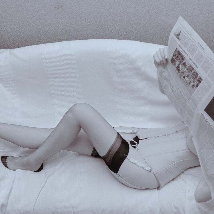 Modell in Korsett mit Strumpfhaltern und Nylons liest liegend eine Zeitung. Das Gesicht ist von der Zeitung verdeckt.