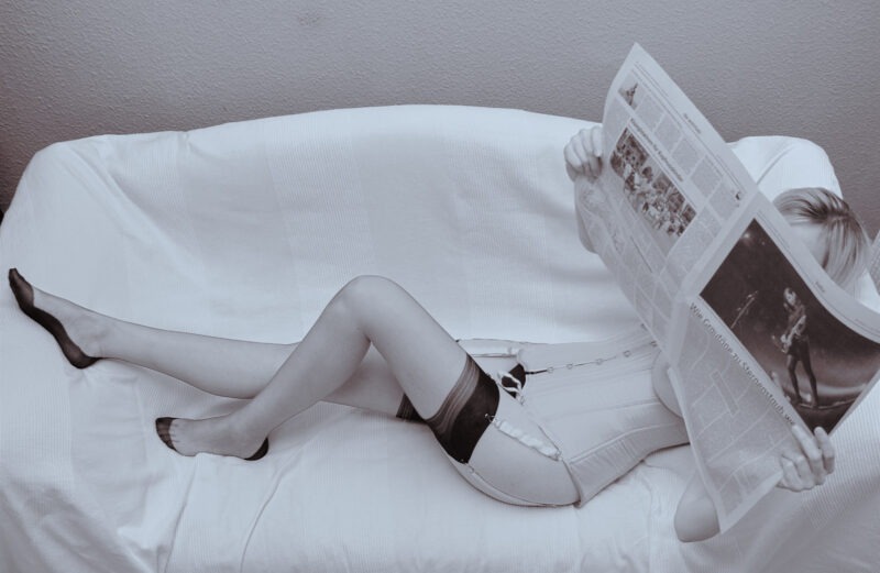 Modell in Korsett mit Strumpfhaltern und Nylons liest liegend eine Zeitung. Das Gesicht ist von der Zeitung verdeckt.