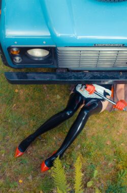 AG Nahtstrumpf 0,2 mm real seamed latex stockings Ein weibliches Modell in schwarzen Latexstrümpfen liegt mit einem Schraubenschlüssel unter einem Land Rover Defender. Sie trägt eine Serviererinnenschürze und rote Lederhandschuhe